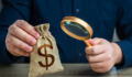 A man examines a dollar money bag through a magnifying glass. Investigating capital origins. Anti money laundering, tax evasion. Study terms and conditions on deposit or loan. Find investments